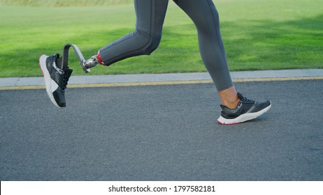 Close Up Of Woman With Prosthetic Leg Running On Park Road. Female Sportsperson Training Outdoors In Slow Motion. Runner Girl Feet Jogging On Asphalt Road