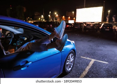 Close up of woman s legs dangling out a car window parked in front of a big white screen at drive in cinema. Concept of carefree leisure time