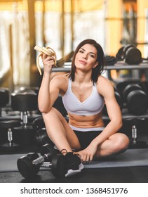 Close Up Of A Woman Is Sitting On A Mat In The Gym And Holding A Banana While Workout Break In A Fitness Gym. Lifestyle And Health.