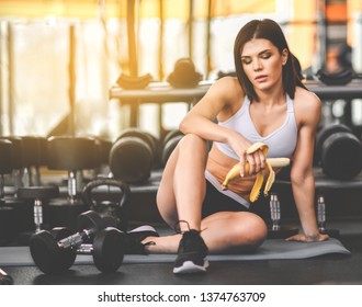 Close Up Of A Woman Is Sitting On A Mat In The Gym And Holding A Banana While Workout Break In A Fitness Gym. Lifestyle And Health.