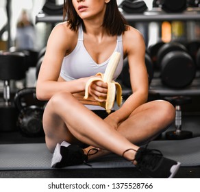 Close Up Of A Woman Is Sitting On A Mat In The Gym And Holding A Banana While Workout Break In A Fitness Gym. Lifestyle And Health.