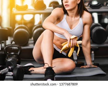 Close Up Of A Woman Is Sitting On A Mat In The Gym And Holding A Banana While Workout Break In A Fitness Gym. Lifestyle And Health.