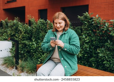 Close up of woman sitting on the modern bench and using smartphone outdoors. Urban lifestyle concept. Happy 30s woman chatting on cell phone - Powered by Shutterstock