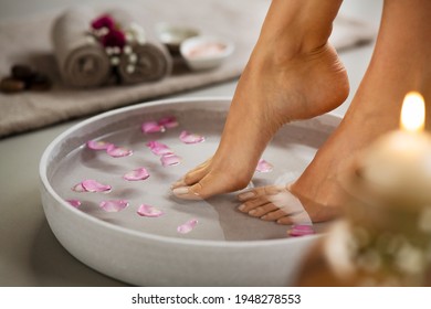 Close up of woman soaking her feet in grey bowl of water with floating petals at beauty luxury spa. Detail of woman feet at wellness center on pedicure procedure. Foot spa and body care concept.