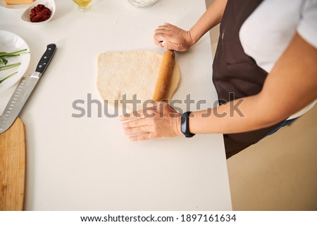 Close up of a woman standing near the kitchen table and rolling dough with a wooden rolling pin