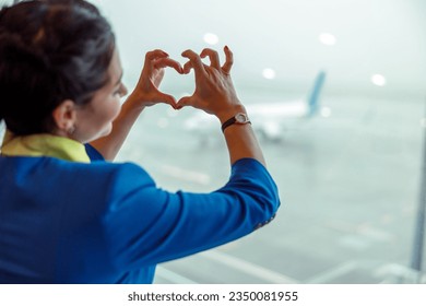 Close up of woman stewardess making heart sign with hands while looking out the window in airport terminal - Powered by Shutterstock