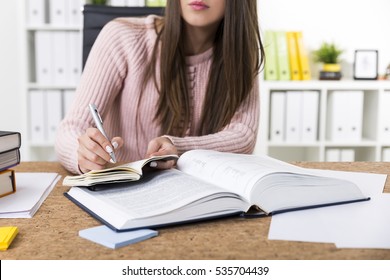 Close Up Of A Woman Wearing A Pink Sweater And Writing Information She Has Found In A Large Book Into Her Notebook