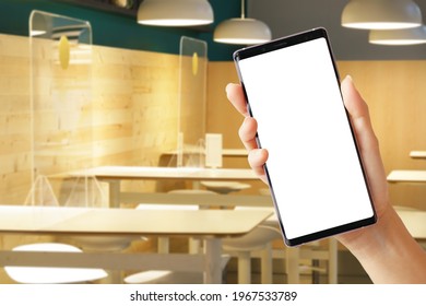 Close Up Of A Woman's Hand Hold A Smartphone With White Blank Screen And Empty Restaurant Seats In Background. Mobile Application, Business, Services, Food Delivery, Apps, Covid 19, Stop Spreading.