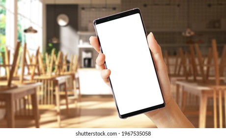 Close Up Of A Woman's Hand Hold A Smartphone With White Blank Screen And 3D Empty Restaurant Seats In Background. Mobile Application, Business, Services, Food Delivery, Apps, Covid 19, Stop Spreading.