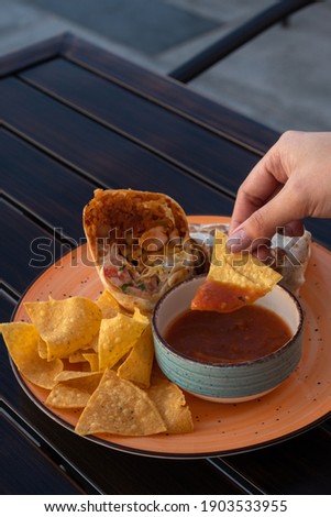 A close up of a woman's hand holding a taco dipped in sauce. A plate of burrito and tacos on an outdoor wooden table.