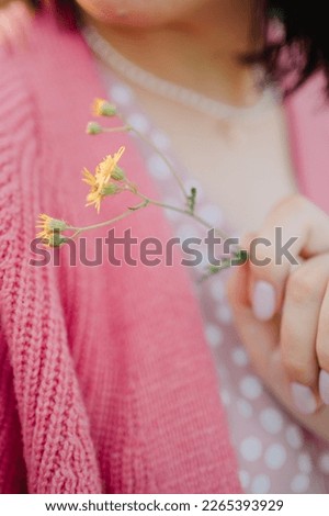 Close up of woman's hand holding yellow wildflower. Pink dress and pink jacket on the background. Vertical.