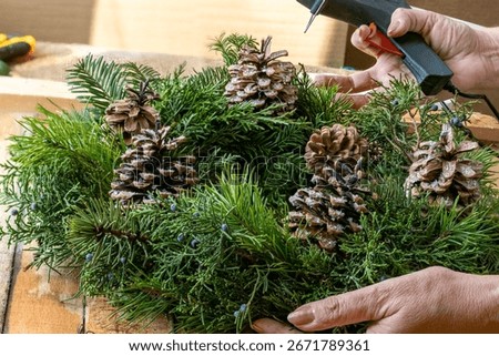 Close up of woman's hands securing pine cones on Christmas wreath using hot glue gun. Making Christmas wreath from natural materials that emit aroma, essential oils and phytoncides.