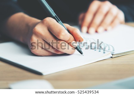 Close up of woman's hands writing in spiral notepad placed on wooden desktop with various items