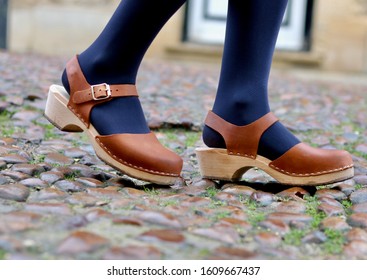 A close up of a woman's legs walking with beautiful leather and wood clog shoes on a cobblestone cobble street
