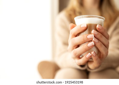 Close up women's hands and fingers with a beautiful French manicure holding cup transparent glass with coffee with white foam.Copy space for text, banner. Beige knitted sweater and background. - Powered by Shutterstock