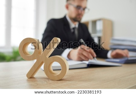 Close up of wooden percent sign on table as symbol of corporate tax and interest rate. Percent sign standing on background of serious busy man in suit calculating on calculator. Blurred background.