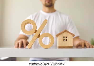 Close up of a wooden percent symbol on a white table with a young man and a little house in the background. Real estate and mortgage interest rates concept - Shutterstock ID 2162398653