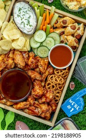 Close Up Wooden Tray Filled With Football Game Day Tailgate Party Food And Snacks With Wings, Wieners, Chips, Dips And Sauces With Football Themed Decorations