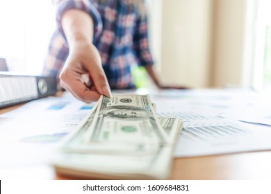 close up of worker picking up pile of cash money bills stack on the wooden table, laying on top of work documentation of the business with folder files nest to it, representing business and payment