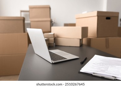 Close up workspace with open laptop, stack of papers on table, multiple cardboard boxes of various sizes in background, no people. On-line ecommerce store, dropshipping business, marketplace website