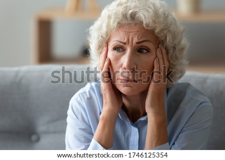 Close up worried elderly 70s woman seated on sofa touch head with hands deep in sad thoughts, memory loss dementia mental problems, chronic senile diseases symptoms, life troubles, loneliness concept