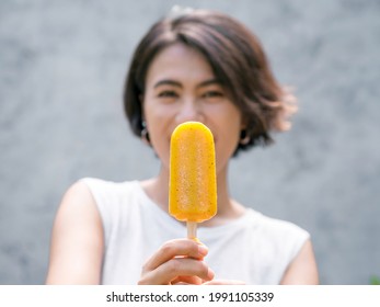 Close up yellow popsicle in beautiful happy Asian woman’s hand wearing casual white sleeveless shirt, outdoors. Woman showing popsicles. Smiling female enjoying ice lolly in summer. - Powered by Shutterstock