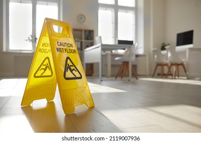 Close Up Of Yellow Standing Plastic Sign With Human Figure That Slips And Falls And Words Caution Wet Floor Placed By Janitorial Service In Empty Clean Sunny Office With Desks, Chairs And Computers