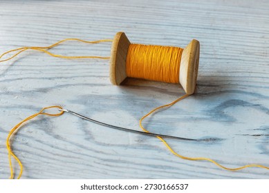 Close up of a yellow thread spool with loose thread and a sewing needle on a light wooden tabletop