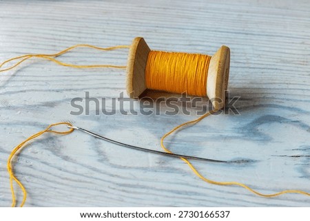 Close up of a yellow thread spool with loose thread and a sewing needle on a light wooden tabletop