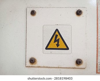 Close up of a yellow triangle high voltage electrical hazard warning sign on a white metal plate with rusty screws
