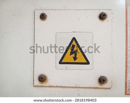 Close up of a yellow triangle high voltage electrical hazard warning sign on a white metal plate with rusty screws