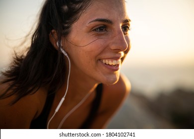 Close up of an young active sporty athlete happy smiling woman is taking a break after making running and jogging workout on a top of rock with seascape at sunset.