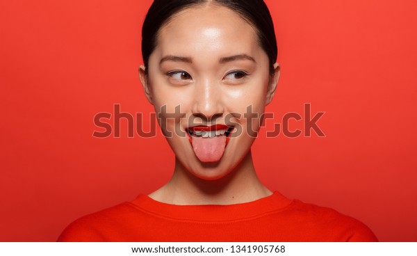 舌を出して目をそらすアジアの若い女性の接写 赤い背景に変な顔をした韓国の女性モデル の写真素材 今すぐ編集