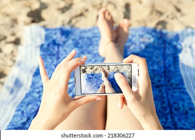 Close up of a young and attractive woman tourist hands together holding a modern technology smartphone and taking a selfie picture of her own legs while laying down on a sandy beach on holiday. - Powered by Shutterstock