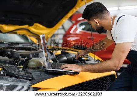 Close up of young bearded engineer mechanic under car hood checking engine with laptop using electronic diagnostic equipment to tune sport yellow vehicle.