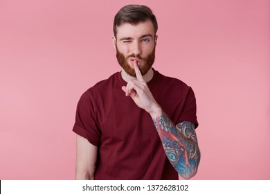 Close up of young bearded man with tattooed hand keeps fore finger on lips, demonstrates silence gesture, isolated over pink background with one rised with raised eyebrow.