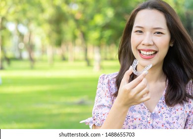 Close Up Young Beautiful Asian Woman Smiling With Hand Holding Dental Aligner Retainer (invisible) At Outdoor Nature Park And Garden Background For Beautiful Teeth And Dental Treatment Course Concept