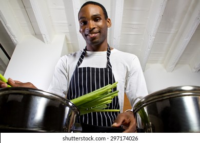 Close Up Of A Young Black Man Wearing An Apron And Cooking At Home. He Is In A Domestic Kitchen And Preparing A Vegetarian Meal.