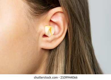 Close Up Of A Young Blonde Woman's Ear With Polyurethane Earplug Isolated On A Gray Background. Anti-noise Earbuds For Sleep, Rest, Travel, Airplane