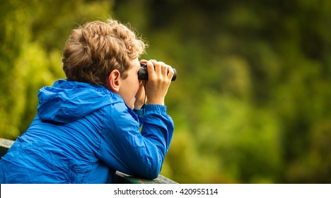 close up of a young boy in a blue coat looking through binoculars at birds with a blurred green background