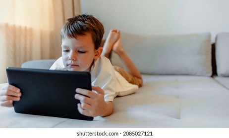 Close up of a young boy playing games on his tablet while laying down on the couch in the living room. Happy boy aged 5 reading an e-book using his tablet. Small boy surfing the net on touchpad.