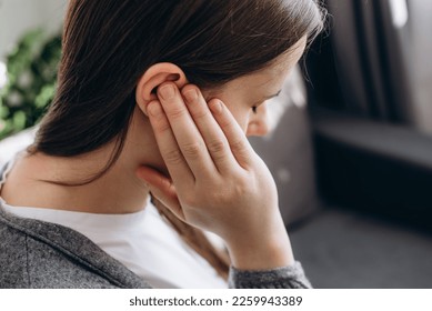 Close up of young brunette female holding painful ear, suddenly feeling strong ache. Unhealthy caucasian woman 20s suffering from painful otitis sitting on couch at home. Health problems concept