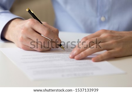 Close up young businessman sitting at table, filling in questionnaire, financial document, bank loan form. Successful male entrepreneur signing contract agreement, putting signature on franchising.