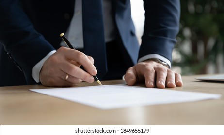 Close up young businessman standing near table with pen in hands, ready signing profitable offer agreement after checking contract terms of conditions, executive manager involved in legal paperwork. - Shutterstock ID 1849655179