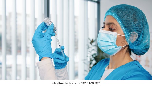 Close Up Of Young Busy Hindu Female Healthcare Specialist Nurse In Protective Uniform And Medical Mask Holding In Hands Ampoule With Coronavirus Vaccine Preparing Injection For Covid Vaccination,