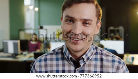 Close up of young caucasian male office worker turning his head and smiling into the camera in the modern office. Working people on the blurred background. Portrait