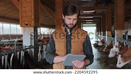 Close up young caucasian man in orange vest using tabletPC and working in farm. Male farmer tapping and scrolling on tablePC in shed. Farming concept.