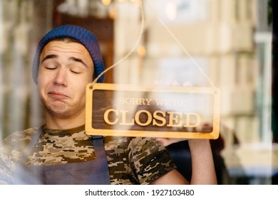 Close Up Of Young Caucasian Man Wearing Blue Cap And Grey Apron About To Close Business In His Own Coffee Shop And Hold A Sign To Close His Own Shop. Funny Face Emotion.