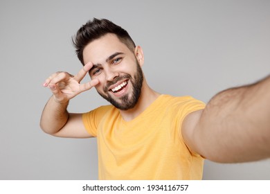 Close up young caucasian smiling happy bearded attractive man in casual yellow basic t-shirt doing selfie shot on mobile phone show victory v-sign gesture isolated on grey background studio portrait