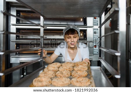 Close up of young caucasian woman baker is holding a tray with fresh bagels and looking to them at baking manufacture factory. Tasty bakery factory concept.